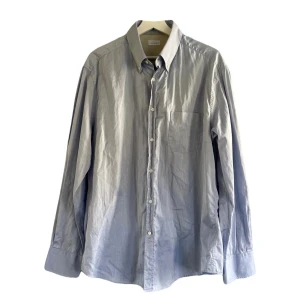 Ljusblå skjorta Brunello Cucinelli - Klassisk ljusblå skjorta från Brunello Cucinelli i 100% bomull. Skjortan har lång ärm, bröstficka och knäppning framtill med vita knappar. Perfekt för dig som gillar stilren och tidlös design med italiensk känsla. Står XXL men passar XL