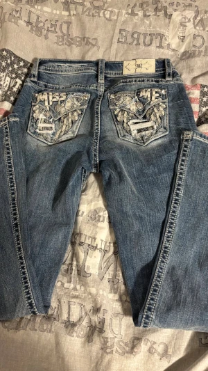 Miss Me blå jeans med broderade fickor - Säljer ett par blå Miss Me jeans med unika bakfickor som har broderade vingar, nitar och slitningar. Fickorna har texten 'MISS ME' och coola detaljer i vitt och silver. Perfekta för dig som gillar statement-plagg. Det är bara att fråga om du vill ha mer bilder, är 170cm för referens💕