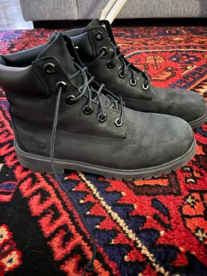 Svarta vattenimpregnerade timberland boots - Säljer ett par klassiska svarta Timberland boots i mocka med robust snörning och vadderad krage. Skorna har rund tå, grov sula och diskret Timberland-logga på sidan. Perfekta för höst och vinter när du vill ha både stil och funktion.