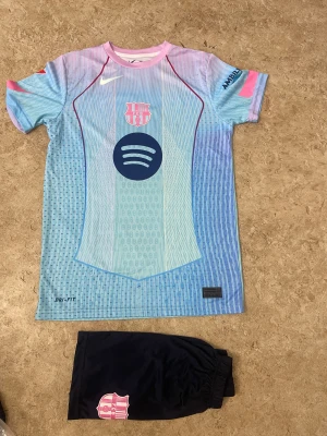 FC Barcelona Lamine Yamal matchställ - Säljer ett FC Barcelona matchställ med Lamine Yamal #10. Tröjan är i ljusblått och rosa med mörkblå detaljer, klubbmärke och sponsorloggor. Shortsen är mörkblå med klubbmärke. Perfekt för fotbollsträning eller att bära som supporter.