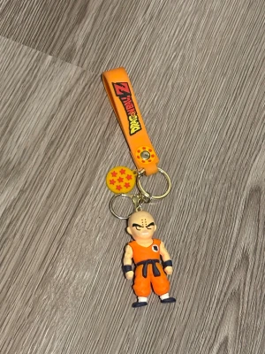 Dragon Ball Z Krillin nyckelring - Nyckelring med Krillin från Dragon Ball Z. Figurerad i orange dräkt med mörkblå detaljer och japanskt tecken på bröstet och ryggen. Nyckelringen har även en orange rem med Dragon Ball Z-logga och en gul drakkula med röda stjärnor. Perfekt för animefans! Dock har den blivit  missfärgad och tappat lite guldfärg.