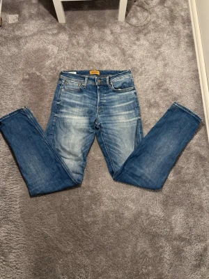 Blå jeans från Jack & Jones, 29/32 - Snygga blå jeans från Jack & Jones med klassisk femficksdesign och slitna detaljer framtill och baktill. Jeansen har raka ben och normal passform, tillverkade i bomull med tydliga kontrastsömmar och läderpatch bak i midjan. Använd ett par gånger inga fläckar dem är i ett bra skick hör av dig om du har några frågor .😊