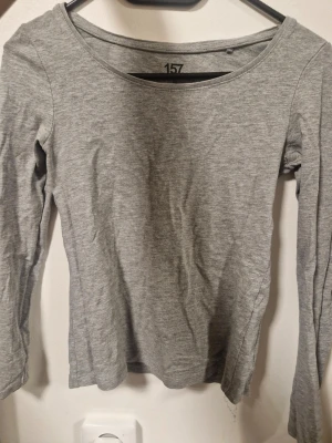 Grå långärmad topp från 157 - En enkel och stilren grå långärmad topp från 157. Modellen har rund halsringning och är figurnära utan synliga detaljer. Perfekt att matcha med jeans eller kjol för en clean look. Materialet känns mjukt och stretchigt, troligen bomull eller polyester.