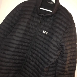 Ny svart pufferjacka från Helly Hansen  - Svart pufferjacka från Helly Hansen i storlek 2XL. Jackan har quiltad design, dragkedja framtill och innerficka. Perfekt för kyliga dagar med sin lätta och smidiga passform. Klassisk HH-logga på bröstet och hög krage för extra värme.