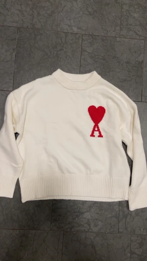 Vit stickad tröja från Ami Paris - Vit stickad tröja från Ami Paris med röd hjärtapplikation och bokstaven A på bröstet. Tröjan har ribbade muddar vid hals, ärmslut och nederkant samt en klassisk rund halsringning. Perfekt för dig som gillar stilrena detaljer och vill sticka ut med en ikonisk design.