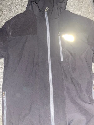 Svart vindjacka från The North Face - Svart vindjacka från The North Face med huva och hel dragkedja framtill. Jackan har flera praktiska fickor med dragkedja och meshpaneler vid axlarna för extra ventilation. Perfekt för dig som gillar att vara aktiv utomhus och vill ha en stilren look.