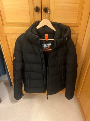 Svart pufferjacka från Superdry - Svart pufferjacka från Superdry med huva och dragkedja. Jackan har sportig look, diskreta logodetaljer i orange och svart samt två fickor med dragkedja. Perfekt för kalla dagar och riktigt skön att bära. Klassisk quiltad design och normal passform.