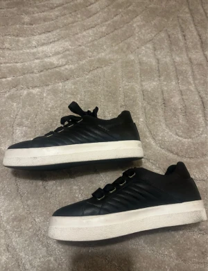 Svarta sneakers från Gant med snörning - Svarta sneakers från Gant med breda svarta snören och guldfärgade öljetter. Skorna har en vit, platt sula och är tillverkade i skinn med diskreta sömmar på sidorna. Perfekt för dig som gillar stilrena och klassiska sneakers. Använda en gång men var för stora därav säljer dem :(