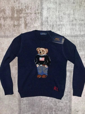 Mörkblå stickad tröja Polo Bear - Mörkblå stickad tröja från Polo Ralph Lauren med ikonisk Polo Bear-motiv på bröstet. Rund hals, långa ärmar och RL-broderi i rött vid nederkanten. Materialmix med merinoull, polyester, akryl och nylon. Perfekt för dig som gillar klassisk streetstyle med en twist.
