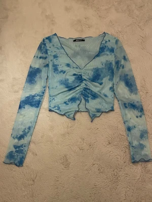 Blå mönstrad meshblus Gina Tricot XS - Snygg meshblus från Gina Tricot i storlek XS. Blusen har ett blått batikmönster, v-ringning och lång ärm. Framtill finns en cool snörning som ger en rynkad effekt och asymmetrisk nederkant. Materialet är tunn mesh som känns lätt och fräsch.