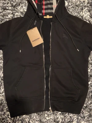 Svart burberry hoodie - Svart hoodie från Burberry i storlek M, tillverkad i 100% bomull. Huva med klassiskt Burberry-    rutigt mönster i svart, vit, grå och röd. Dragkedja framtill, snöringen vid huva och två fickor. Perfekt för dig som gillar stilrena streetwear-vibbar.