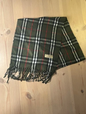 Grön rutig Burberry ullväst - Snygg grön väst från Burberry med klassiskt rutigt mönster i svart, vitt och rött. Västen har fransar nedtill och är tillverkad i mjuk ull. Perfekt för dig som vill ha en cool och tidlös look med designer-touch.