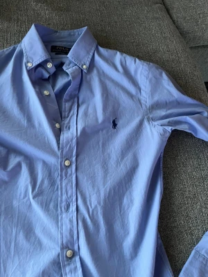 Blå skjorta Polo Ralph Lauren XS - Snygg ljusblå skjorta från Polo Ralph Lauren i slim fit-modell. Klassisk button down-krage, vita knappar och ikoniska broderade loggan på bröstet. Skjortan är långärmad och tillverkad i bomull, perfekt för en clean och stilren look.