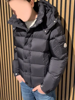 Moncler vezere jacka - Säljer en svart pufferjacka från Moncler med avtagbar huva och klassisk logga på ärmen. Jackan är quiltad och dragkedja samt knappar framtill . Perfekt för kalla dagar och riktigt snygg till jeans.