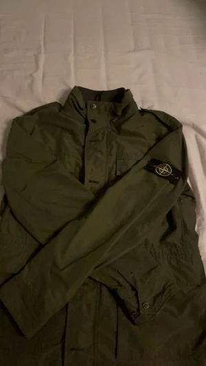 Grön vindjacka från Stone Island - Säljer en grön vindjacka från Stone Island med klassisk badge på vänster ärm. Jackan har hög krage, två stora fickor framtill och både dragkedja och knappar. Materialet är slitstark polyester och passformen är normal. Perfekt för dig som vill ha en clean och stilren look.