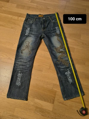 Unika jeans med drakmotiv och broderier - Säljer ett par coola blå jeans från DAKS med slitna detaljer, broderier och ett stort drakmotiv på ena benet. Jeansen har raka ben, fem fickor och kontrastsömmar. Bakfickorna har broderad DAKS-logga. Perfekt för dig som vill sticka ut med din stil.