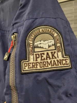 Peak Performance Klondyk PA blå parkas jacka, storlek S passar M - Säljer en marinblå parkas från Peak Performance med snygga detaljer som patch på ärmen, röda dragskor och flera praktiska fickor med dragkedja. Jackan har hög krage, huva och är perfekt för kalla dagar. Materialet är tåligt och jackan har en clean outdoor-look.