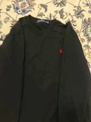 Svart sweatshirt från Polo Ralph Lauren - Svart sweatshirt från Polo Ralph Lauren med klassisk rund hals och lång ärm. Tröjan har det ikoniska röda Polo-logot broderat på bröstet och är gjord i mjuk bomull. Perfekt för dig som gillar stilrena och tidlösa plagg.