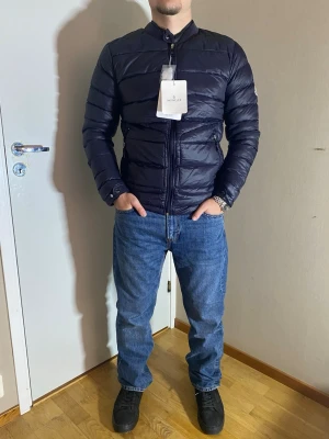 Moncler dunjacka  - Säljer en mörkblå, lätt och stilren dunjacka från Moncler. Jackan har quiltad design, hög krage och dragkedja framtill samt två sidofickor med dragkedja. Insidan har Monclers klassiska serietidningsmönster. Perfekt för kyliga dagar och riktigt snygg passform.