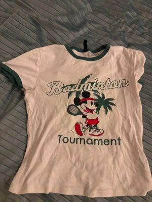 Vit t-shirt med Mickey Mouse och badminton - Vit t-shirt med gröna detaljer på ärmslut och krage. Tryck framtill med Mickey Mouse i sportkläder, palmträd och texten 'Badminton Tournament'. Perfekt för dig som gillar sportiga och lekfulla plagg.