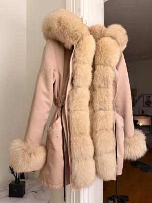 Beige pälsjacka med fluffig huva - Säljer en beige pälsjacka med stor fluffig huva och breda pälsdetaljer längs krage, ärmslut och framkant. Jackan har en snygg passform och band i midjan. Perfekt till vintern.