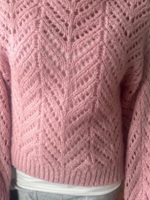 Rosa stickad tröja med hålmönster - Säljer en superfin rosa stickad tröja med snyggt hålmönster och breda ribbade muddar. Tröjan har lång ärm och en mysig, lite loose passform som ger en avslappnad vibe. Perfekt för dig som vill ha något både gulligt och trendigt i garderoben.