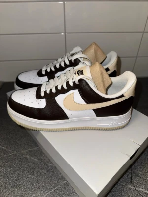 Nike Air Force 1 i brunt och beige - Nike Air Force 1 sneakers i en clean färgkombo med vit tå och sidor, mörkbruna paneler och beige swoosh. Klassisk låg modell med snörning, perforerad tå och vit sula. Ovandel i skinn och detaljer i beige på plös och häl. Perfekta för dig som gillar stilrena sneakers med twist. Skorna är i nyskick!!!