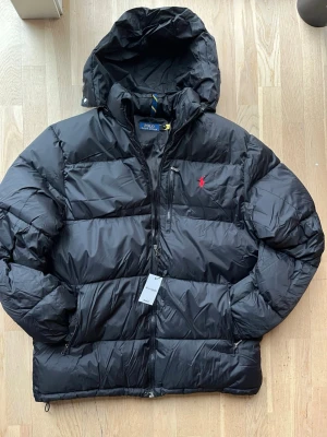 Svart dunjacka från Polo Ralph Lauren - Svart dunjacka från Polo Ralph Lauren med huva, dragkedja och flera fickor med dragkedja. Jackan har en röd broderad logga på bröstet och är quiltad för extra värme. Perfekt för kalla dagar och har en klassisk, sportig look.