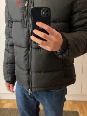 Ralph Lauren pufferjacket  - Svart pufferjacka från Ralph Lauren med röd broderad logga på bröstet. Jackan har huva, flera dragkedjeförsedda fickor och RL-detalj på dragkedjan. Perfekt för kalla dagar och har en klassisk, sportig look.