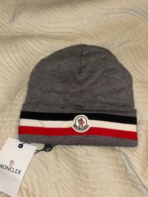 Grå Moncler mössa med ränder - Snygg grå Moncler mössa med klassiska svarta, vita och röda ränder nertill. Mössan har Moncler-logga framtill och är tillverkad i mjukt stickat material. Perfekt accessoar för att hålla stilen under kyliga dagar.Aktiv 24/7✅ Skickas inom 24h📦🚚 Kan gå ner i hugget 😃✅ Hör av er via frågor⁉️