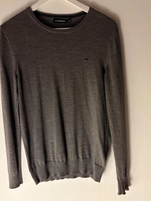 Jlindeberg tröja  - Snygg grå stickad tröja från J.Lindeberg med diskret logga på bröstet. Tröjan har rund hals, ribbade muddar och lång ärm. Perfekt för dig som gillar stilrena och klassiska plagg med en modern touch.