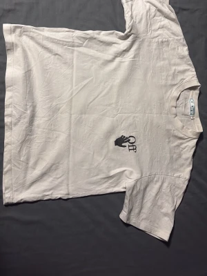 Vit Off-White t-shirt med stor logga - Vit t-shirt från Off-White i bomull med klassisk rund hals och korta ärmar. Liten svart hand-logga framtill och stor grafisk Off-White-logga i blått och svart på ryggen. Snygg och stilren streetwear-vibe.