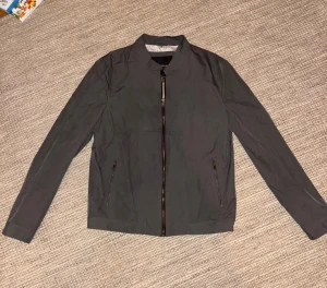 Grå frield jacket från Bruun & Stengade - Snygg grå fieldjacket från Bruun & Stengade i storlek M. Jackan har stilren design med ståkrage, hel dragkedja framtill och två fickor med dragkedja. Insidan har ett diskret mönstrat foder som ger extra vibe. Perfekt för dig som gillar stilren och riktigt snygg jacka för vår/höst/sensommar. Nypris 1100.