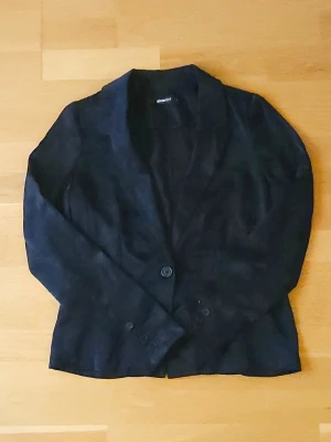 Svart kavaj - Svart kavaj från Gina Tricot med klassisk krage och två knappar framtill. Materialet har en lätt glansig finish och kavajen har långa ärmar med fyra dekorativa knappar vid ärmslutet. Snygg, stilren och passar perfekt att slänga på sig över nyårsoutfiten.