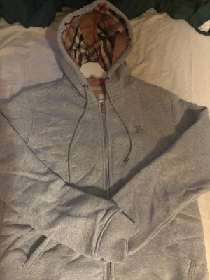 Grå Burberry hoodie - Säljer en grå hoodie från Burberry i storlek M. Tröjan har dragkedja, snörning vid huvan och Burberrys klassiska rutiga mönster på insidan av huvan. Broderad logga på bröstet och två fickor framtill. Perfekt för dig som vill ha en stilren och lyxig look. Passar även S