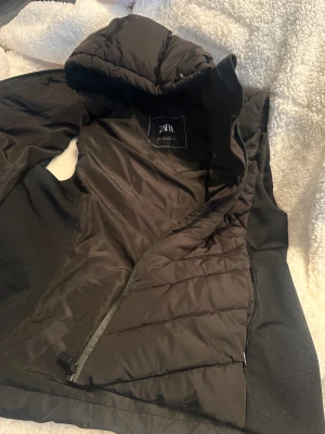 Svart pufferjacka från Zara med huva - Svart pufferjacka från Zara i storlek L. Jackan har en snygg quiltad design, huva och dragkedja framtill. Materialet är polyester och jackan har en normal passform. Perfekt för kalla dagar och enkel att matcha med din stil. Obs helt ny aldrig använda men har fått en liten skada som finns bifogat i en av bilderna storleken L men passar mig som har s