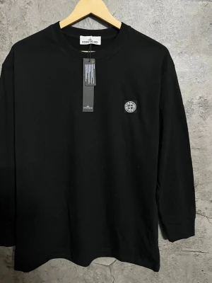 Svart långärmad topp från Stone Island - Svart långärmad topp från Stone Island med rund halsringning och ikonisk kompasslogga broderad på bröstet. Tillverkad i mjuk bomull för en clean och stilren look. Perfekt för dig som gillar streetwear och vill ha något enkelt men ändå med tydlig branding.