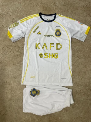 Al Nassr Ronaldo 7 matchställ Adidas - Säljer ett Al Nassr matchställ med Ronaldo 7-tryck. Tröjan är vit med gula detaljer, klubbmärke och sponsorloggor framtill, samt Adidas-logga. Shortsen är vita med klubbmärke. Perfekt för fotbollsfans som vill sticka ut på planen.