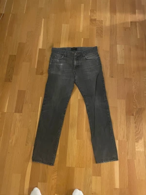 Grå jeansbyxor Tiger of Sweden herr - Snygga grå jeansbyxor från Tiger of Sweden med klassisk femficksdesign och raka ben. Jeansen har diskreta slitningar framtill och svarta detaljer på bakfickorna. Passar dig som gillar stilrena och tidlösa jeans med en modern touch. Börjar bli slitna i grenen men går lätt att laga