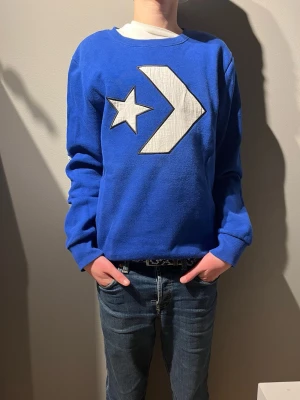 Blå Converse sweatshirt XL - Blå sweatshirt från Converse i storlek XL med stort vitt Converse-logotryck på bröstet. Tröjan har rund hals, långa ärmar och ribbade muddar vid ärmslut och nederkant. Materialet är mjuk bomull och polyester, perfekt för chill dagar.