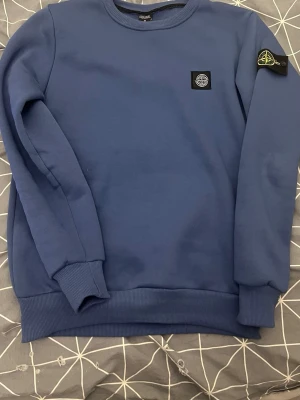 Blå Stone Island sweatshirt - Använd bara ett få tal gånger 4-5 , inga defekter alls, ser Helt ny ut, Säljer en blå crewneck sweatshirt från Stone Island med klassisk patch på vänster ärm och logga på bröstet. Tröjan har rund hals, ribbade muddar och är tillverkad i mjuk bomull. Perfekt för dig som gillar streetwear och vill ha en clean look.