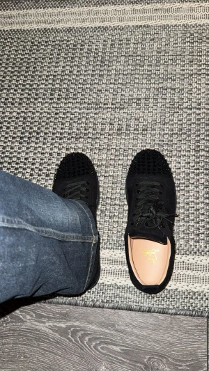 Svarta sneakers med nitar från Louboutin - Säljer ett par svarta sneakers från Louboutin med coola svarta nitar på tån. Skorna har snörning och är tillverkade i mocka med rund tå och platt sula. Perfekt för dig som vill sticka ut med en edgy stil.