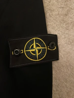 Svart Stone Island sweatshirt - Svart sweatshirt från Stone Island med klassisk rund hals och ikoniskt märke på vänster ärm. Tröjan har en stilren look och är tillverkad i mjuk bomull, perfekt för en clean och avslappnad stil.