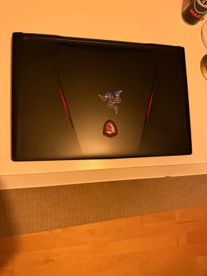 MSI Gaming Laptop - Kraftfull MSI gaming laptop med distinkt design och röda detaljer. Locket har MSI och Razer-klistermärken. Några mindre ytliga repor syns, men överlag gott skick. Perfekt för gaming och krävande uppgifter. Undersidan visar god ventilation för optimal kylning.