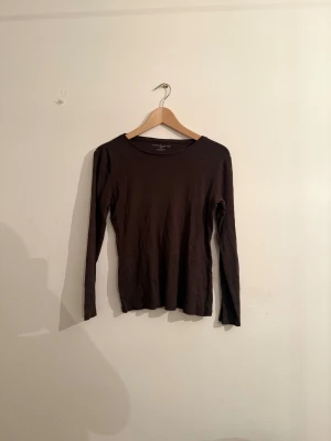 Mörkbrun långärmad topp Ann Taylor - Säljer en enkel och stilren mörkbrun långärmad topp från Ann Taylor i storlek M. Toppen är tillverkad i 100% bomull och har en rund halsringning. Perfekt att matcha med jeans eller kjol för en clean look.