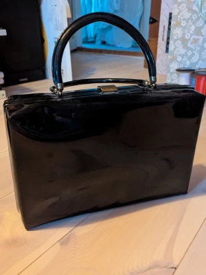 Svart blank handväska med röd insida - Stilren svart handväska i blankt material med klassisk rektangulär form och guldfärgat spänne. Väskan har ett stadigt handtag och insidan är fodrad i rött tyg med ett fack som stängs med dragkedja. Perfekt för dig som gillar retrovibbar och vill sticka ut.