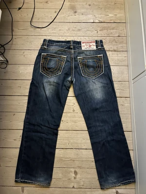 True Religion blå jeans med kontrastsömmar - Säljer ett par blå jeans från True Religion med tydliga kontrastsömmar och ikoniska bakfickor med broderad hästsko. Jeansen har raka ben och guldiga knappar samt detaljerade fickor fram och bak. Klassisk denim-look med slitningar och markerad tvätt. dom är i bra skick. mått: midja rakt över 44cm, ytterbenslängd: 96! dessa är kill jeans men passar såklart alla kön! pris kan diskuteras! 