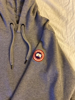 Äkta grå Canada Goose-hoodie med broderad patch. - Äkta Canada Goose-hoodie i grått med klassisk arm-patch. Väldigt mjuk, tjock och i toppskick – endast använd några gånger. Inga defekter alls. Herr modell stl M.   Ordinariepris 3599kr Nypris: 2500kr