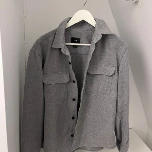 Grå overshirt från H&M - Snygg grå overshirt från H&M med klassisk krage och svarta knappar framtill. Två stora bröstfickor med lock ger en cool och praktisk look. Materialet är mjukt och känns som bomull, perfekt att ha över en t-shirt. Passformen är normal och ärmarna är långa.