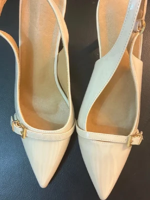 Beige pumps med spetsig tå och klack - Snygga beige pumps med spetsig tå och smal klack. Skorna har en elegant rem med guldfärgat spänne över foten och öppen häl. Tillverkade i blankt syntetmaterial som ger en lyxig känsla. Perfekta för dig som gillar stilrena och trendiga skor.
